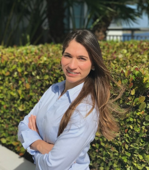 A woman stands confidently with her arms crossed, smiling at the camera, against a backdrop of a well-maintained hedge and palm trees.