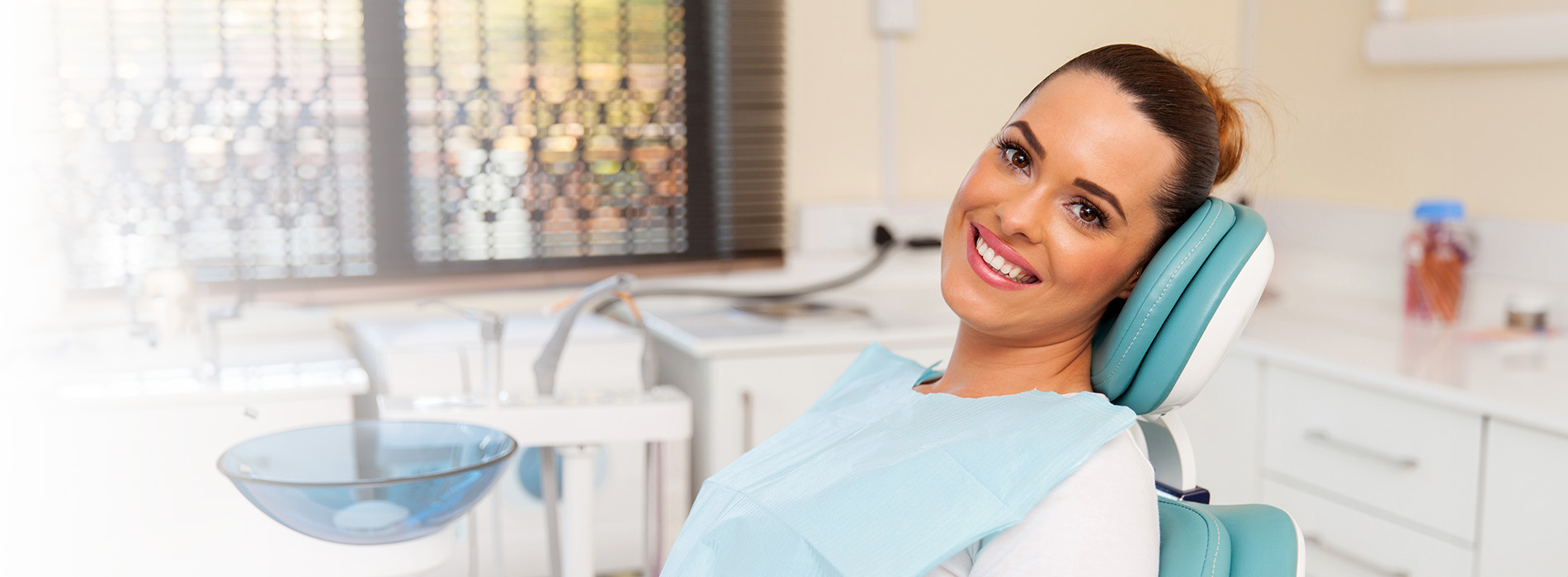 The image shows a smiling woman sitting in a dental chair with her eyes closed, wearing a blue surgical gown and cap, while a dental practitioner is attending to her.