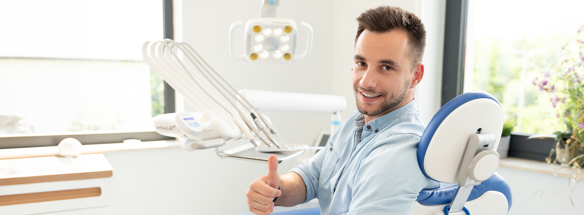 A man sitting at a desk with a dental chair in the background, smiling and giving a thumbs-up gesture.